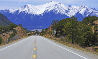 A beautiful shot of the open road with trees on either side of the road and snow-capped mountains in the distance.
