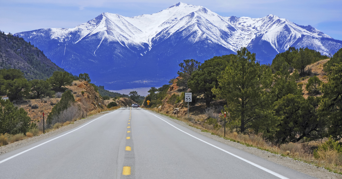 A beautiful shot of the open road with trees on either side of the road and snow-capped mountains in the distance.