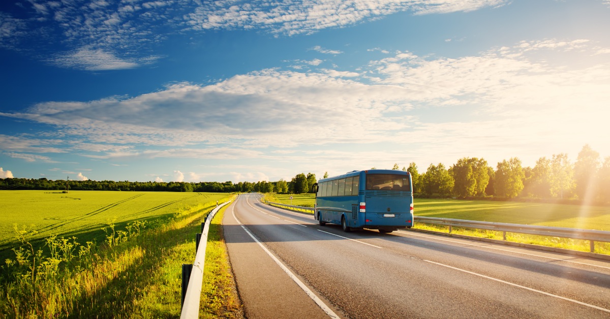 A blue charter bus on a stretch of road with grass on either side and blue skies above.